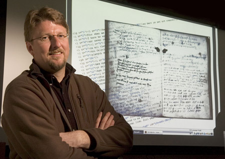 A white man with short hair, facial hair, and glasses wearing a black shirt with brown jacket smiles and crosses his arms in front of a projections of digitized historic documents and computational analysis.