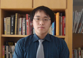 Man with glasses wearing a blue shirt and gray tie with a bookcase behind him.